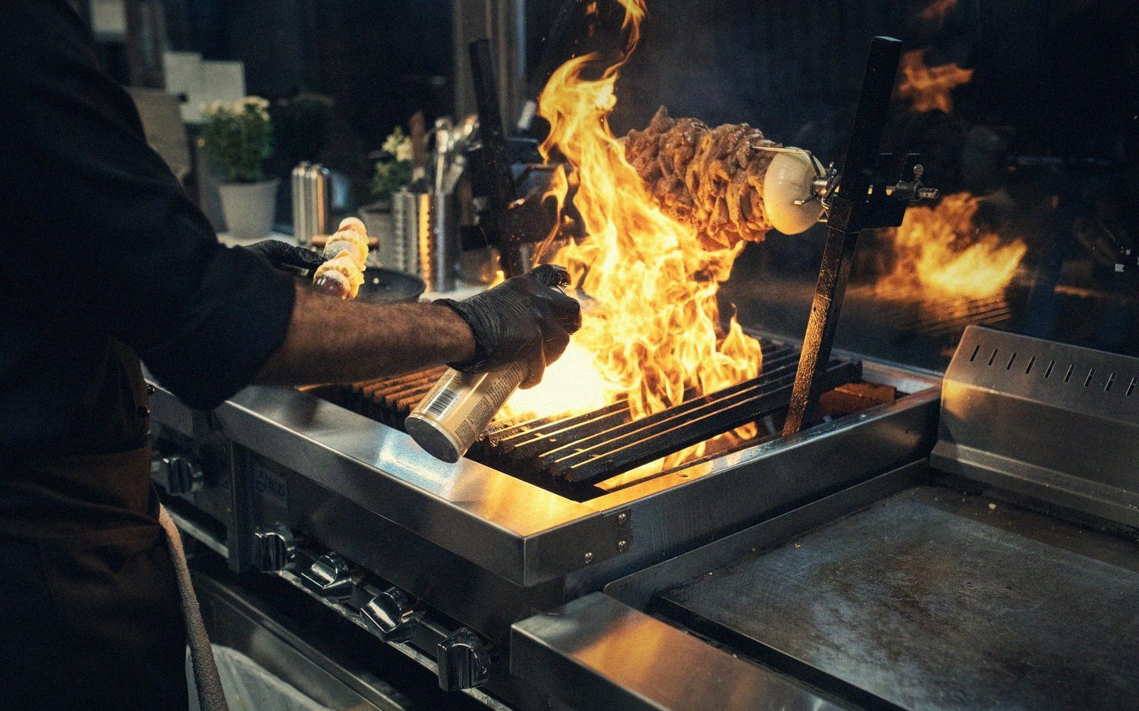 A chef grills meat on a vertical skewer over open flames on a commercial grill