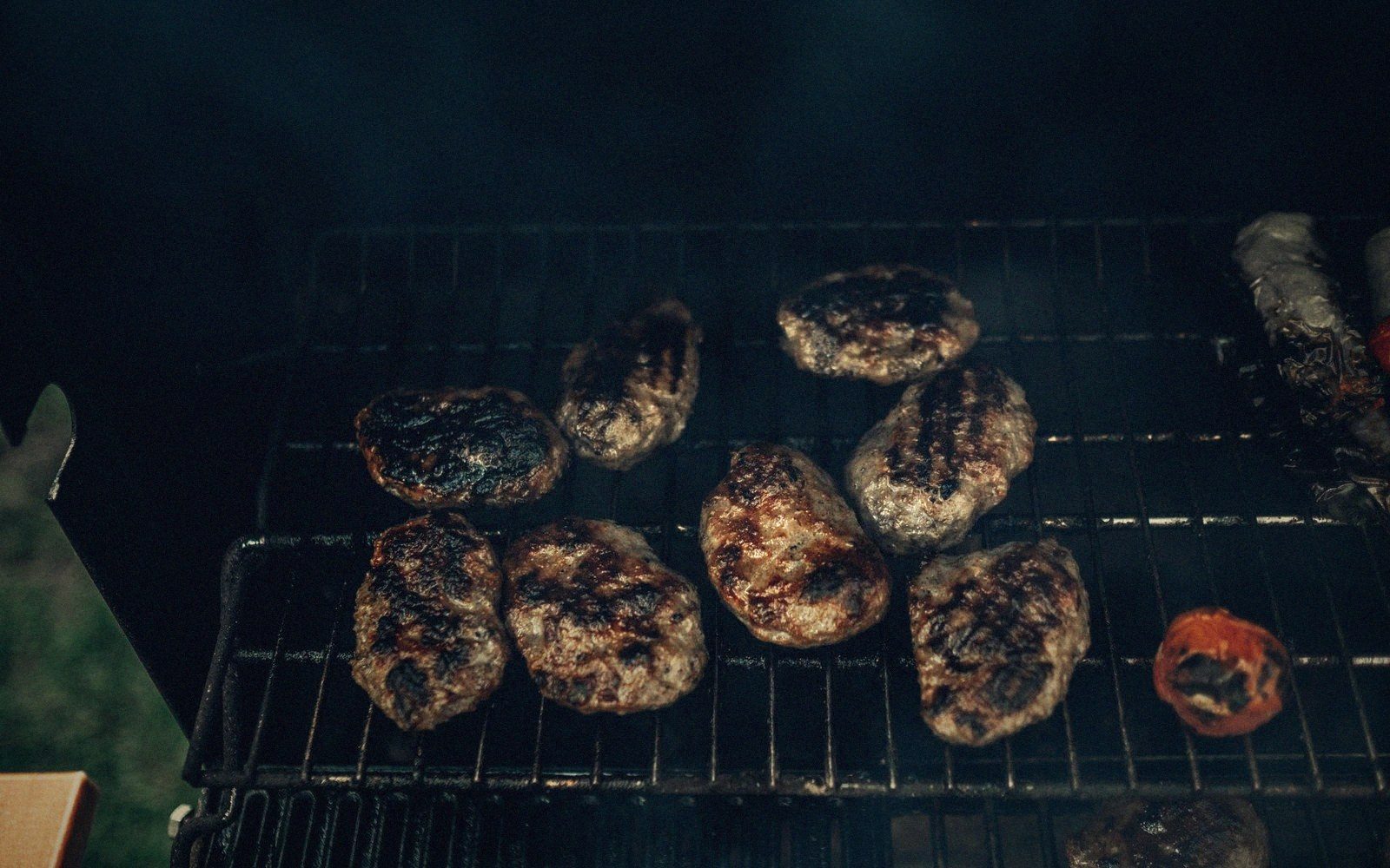 Grilled meat patties and skewers smoking on a charcoal barbecue grate at night