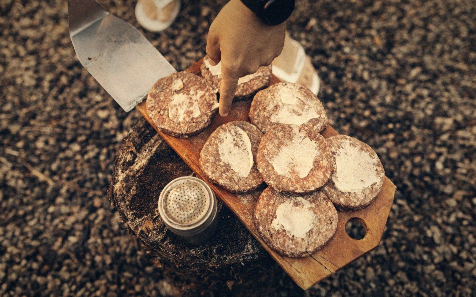 A hand reaches toward raw beef patties dusted with flour on a wooden board