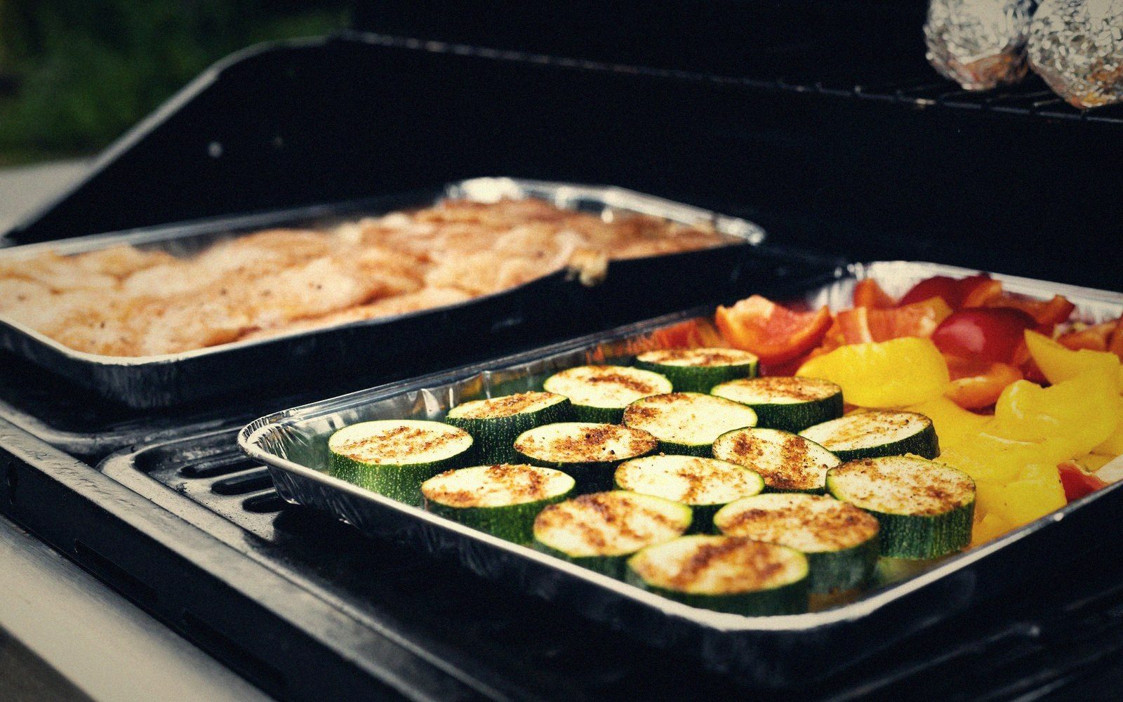 Zucchini, peppers and chicken cook in foil trays on an outdoor grill