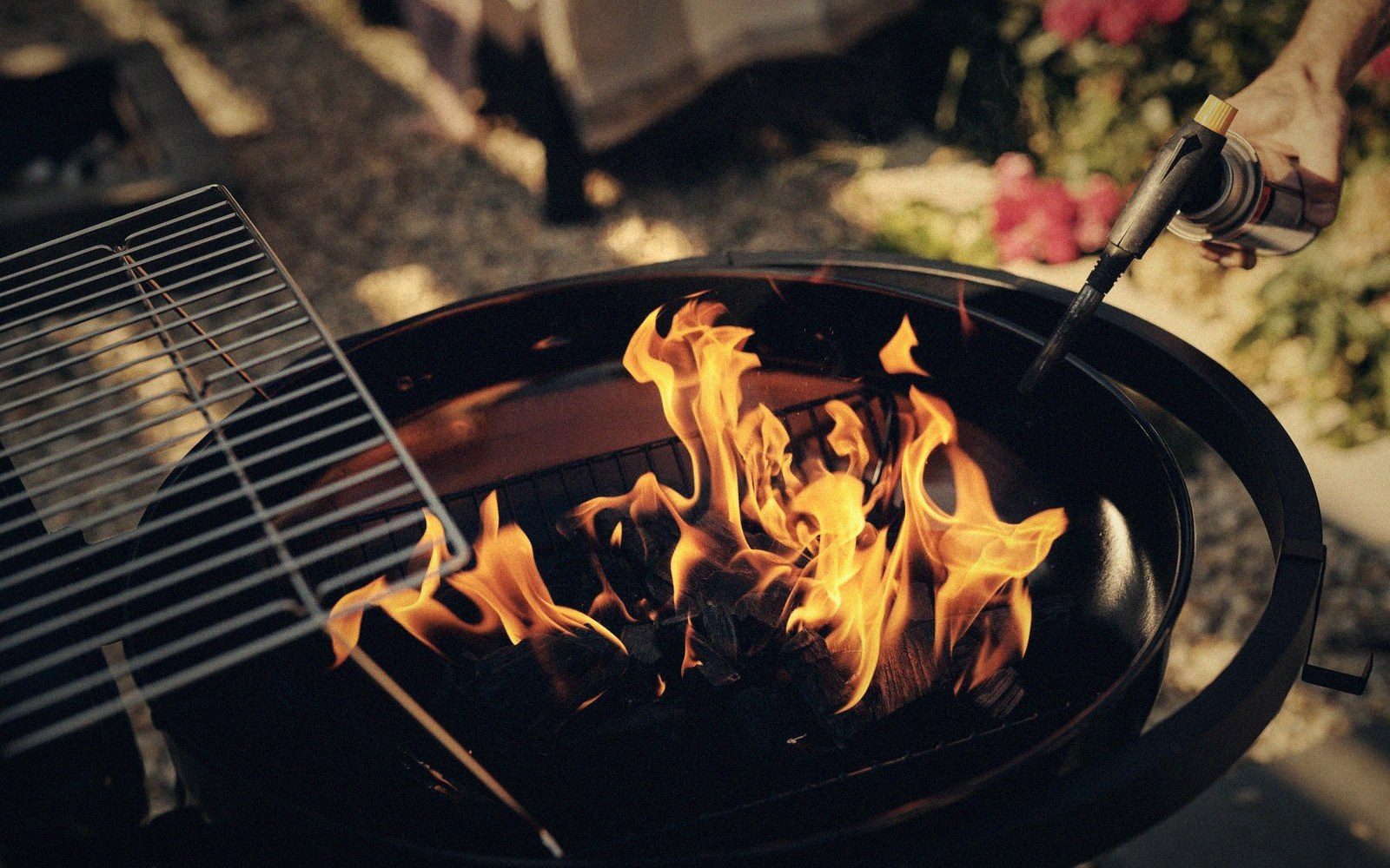 A hand uses a torch to ignite charcoal in a kettle grill with the grate lifted aside