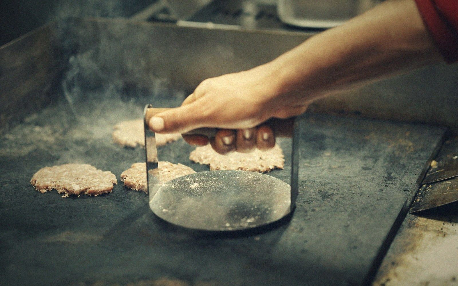 A hand presses a burger patty with a metal weight on a hot griddle