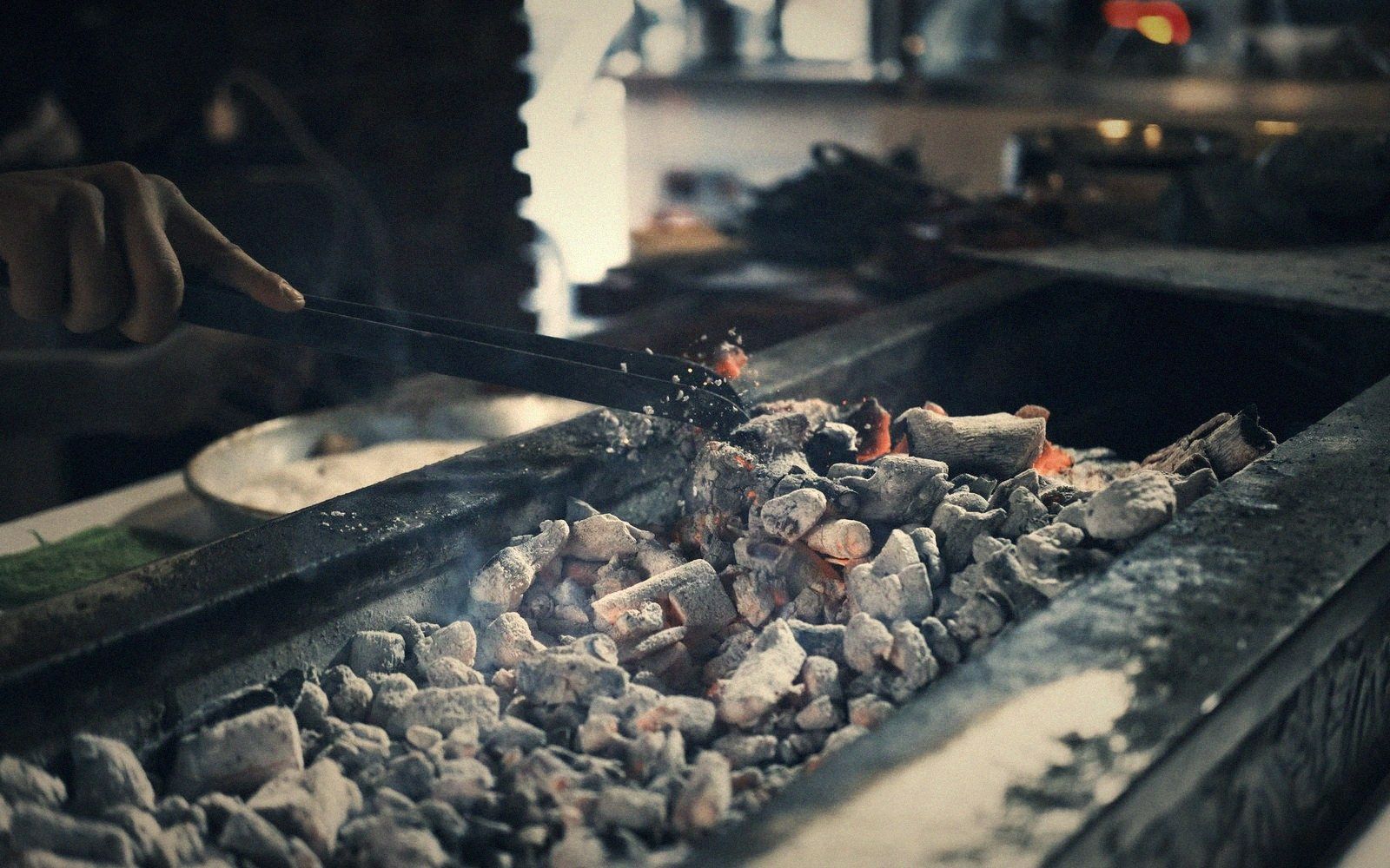 A hand uses tongs to arrange glowing charcoal covered in white ash
