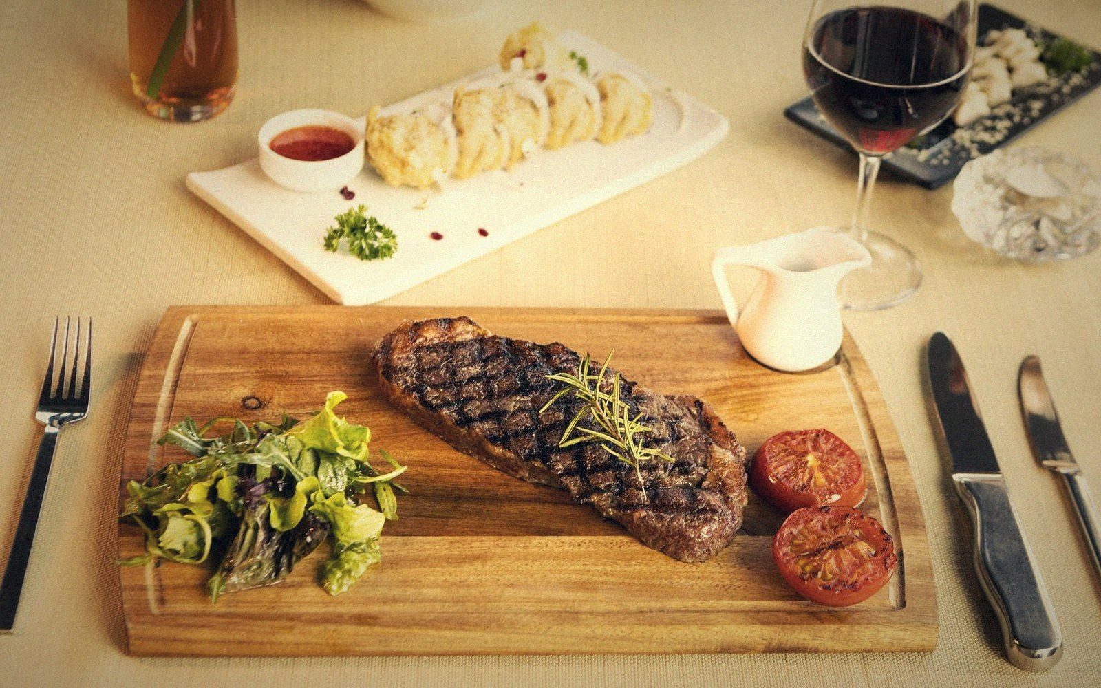 Grilled steak with roasted tomatoes and salad on a wooden board beside wine and dumplings