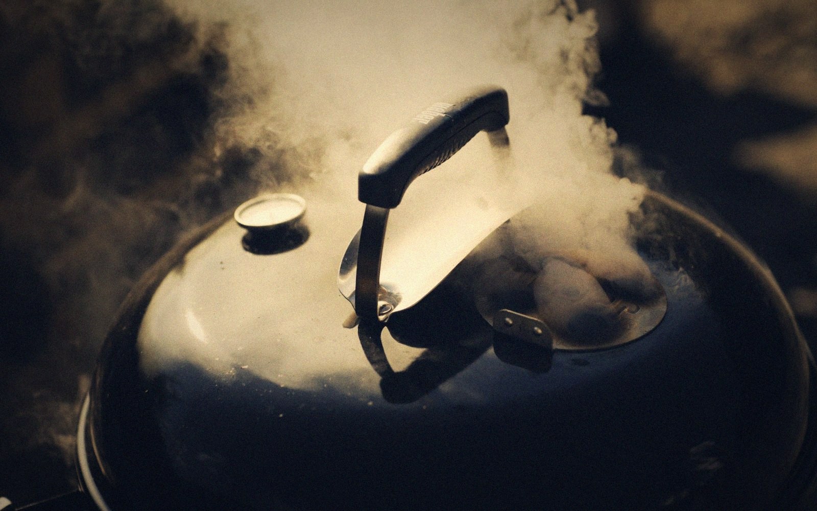 Smoke billows from the vents of a closed kettle grill lid
