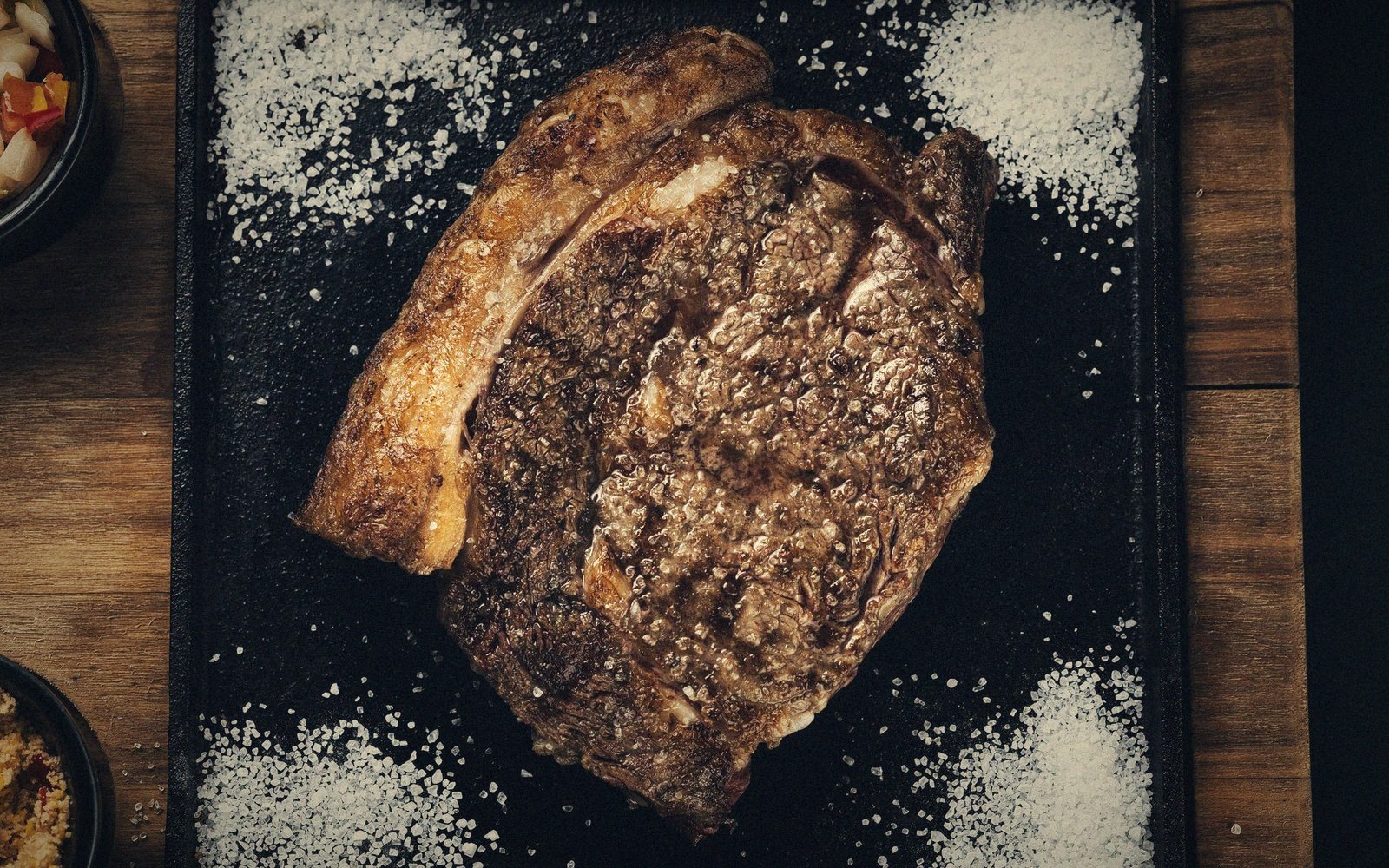 Seared crusted steak with a coarse crust rests on a black board scattered with salt